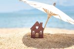 Miniature house and umbrella on beach, blue sea and sky on blurred background. Concept for: If you own property in multiple states you will need to consider it when estate planning.
